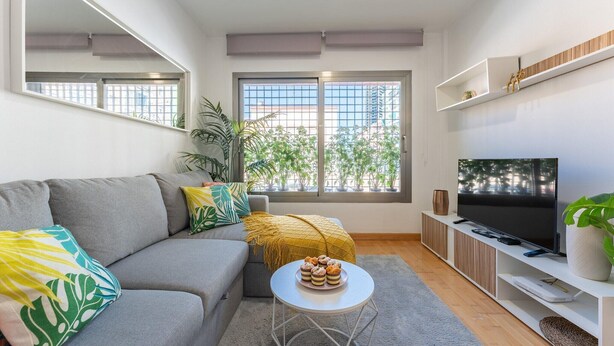 Ein modernes Wohnzimmer mit einem grauen Sofa, einem kleinen weißen Tisch mit Gebäck, einem Fernseher auf einer weißen Ablage und einem großen Fenster mit Blick auf Grünflächen und Gebäude.