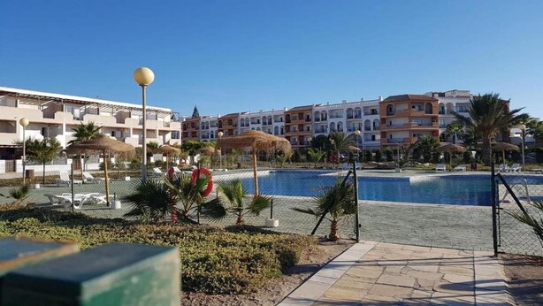 Un área de piscina en un resort, con cielo azul despejado, palmeras y un edificio con balcones al fondo.