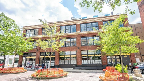 A multi-story building with a brick facade, large windows, and a courtyard with orange seating areas and trees. - Downtown Cincinnati 2BR Suite | Walk to Stadiums (Cincinnati)