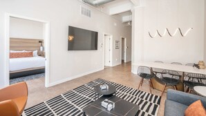 A modern hotel room with a bed, a dining area with chairs and a table, and a large flat-screen TV mounted on the wall. - Downtown Cincinnati 2BR Suite | Walk to Stadiums (Cincinnati)