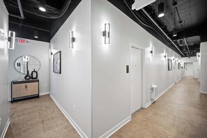 A hallway with white walls, a wooden floor, and a black ceiling with exposed pipes. There are light fixtures on the walls and an exit sign.