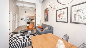 A modern living room with a grey sofa, orange chair, and a wooden table. There are abstract paintings on the wall and a kitchen area in the background. - The 333 Suites 202, Downtown Cincinnati, Sleeps 6 (Cincinnati)
