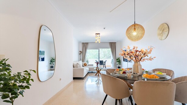 A modern living room with a dining area, featuring a round table, chairs, a large mirror, and a vase with flowers. - Cosy holiday apartment in Elviria, Marbella (Malaga)
