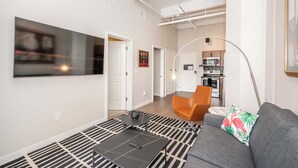 Una sala de estar moderna con un sofá, una mesa de centro y un sillón. Se aprecia una cocina al fondo con electrodomésticos y una zona de comedor.