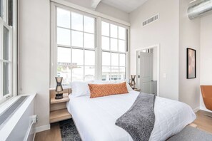A modern bedroom with a large bed, a window, a chair, and a door.