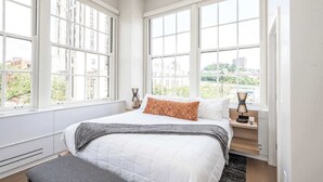 Camera da letto con un letto grande, due comodini con lampade, una panca e vista sugli edifici attraverso ampie finestre.