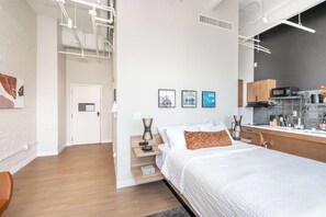 A modern bedroom with a large bed, bedside tables, a nightstand, a desk, and a kitchenette area.