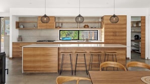 A modern kitchen with wooden cabinetry, a central island with stools, pendant lighting, and a large window with a view.