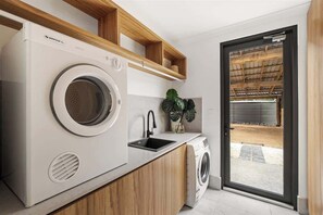A compact laundry area with a front-loading washer and dryer, a sink, wooden shelving, and a view of an outdoor area through a glass door.