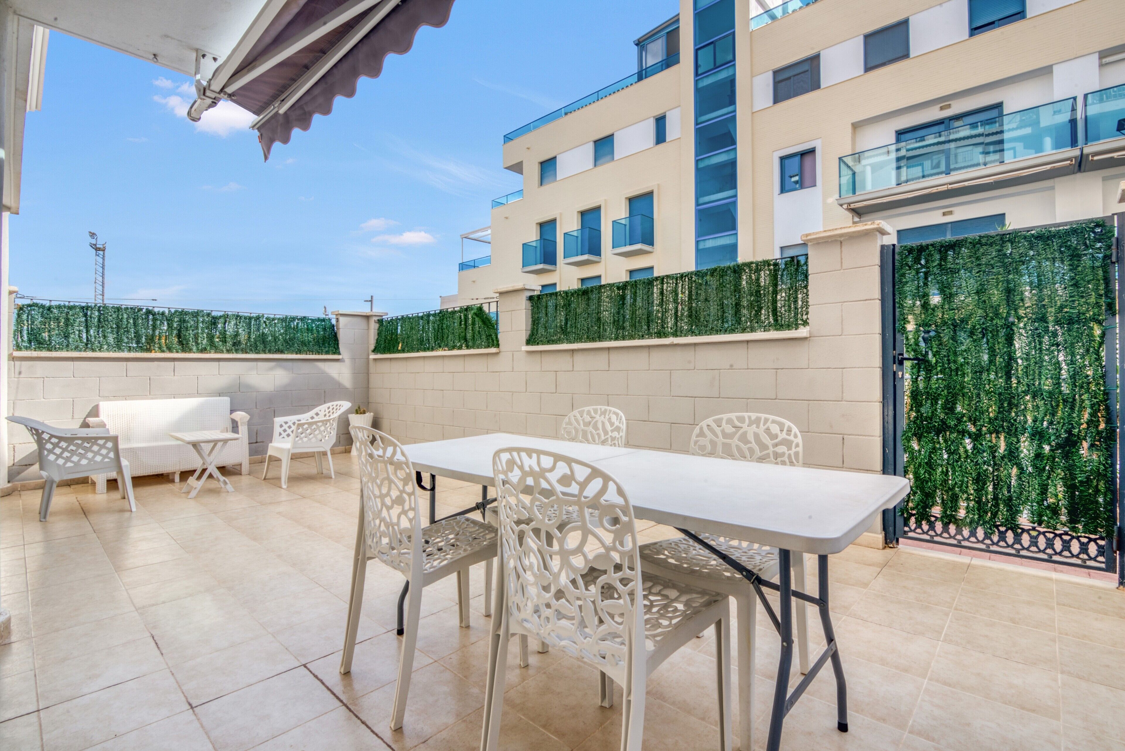 Un patio al aire libre con una mesa y sillas, piso de baldosas y un edificio con balcones y una pared verde al fondo.