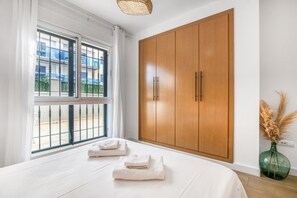 A bedroom with a large wardrobe, a bed with white linens, a window with a view of buildings, and a vase with dried plants. - Apartment 'Prendisole Denia' with Mountain Views, Wi-Fi and Air Conditioning (Denia)