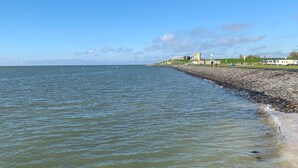 A coastal view with a rocky shoreline, a clear sky, and a few buildings in the distance. - Vacation home Wattglück Tossens Butjadingen - Close to the beach (Butjadingen)