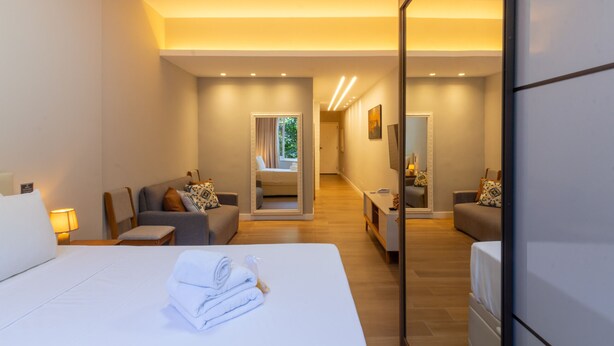 A modern hotel room with a large bed, a sitting area with a sofa and armchairs, a small table, and a mirror.