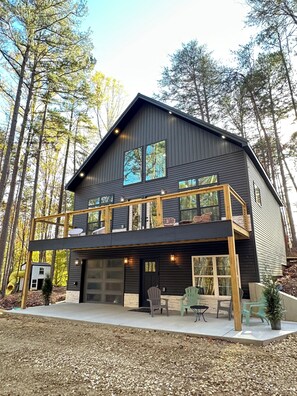 Una casa moderna de dos pisos con fachada oscura, amplias ventanas y una terraza de madera con mobiliario exterior.