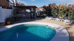 Una piscina con sillones de descanso, un patio con mesas y sillas, y vista a un edificio con tejado de baldosas.