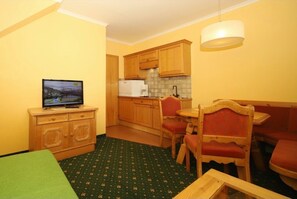 A compact living space with a kitchenette, a dining area with a red chair, a television on a wooden cabinet, and a green rug.