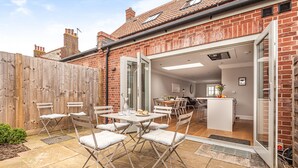 A patio with a table and chairs, a wooden fence, and a brick building with an open door leading to an interior space.