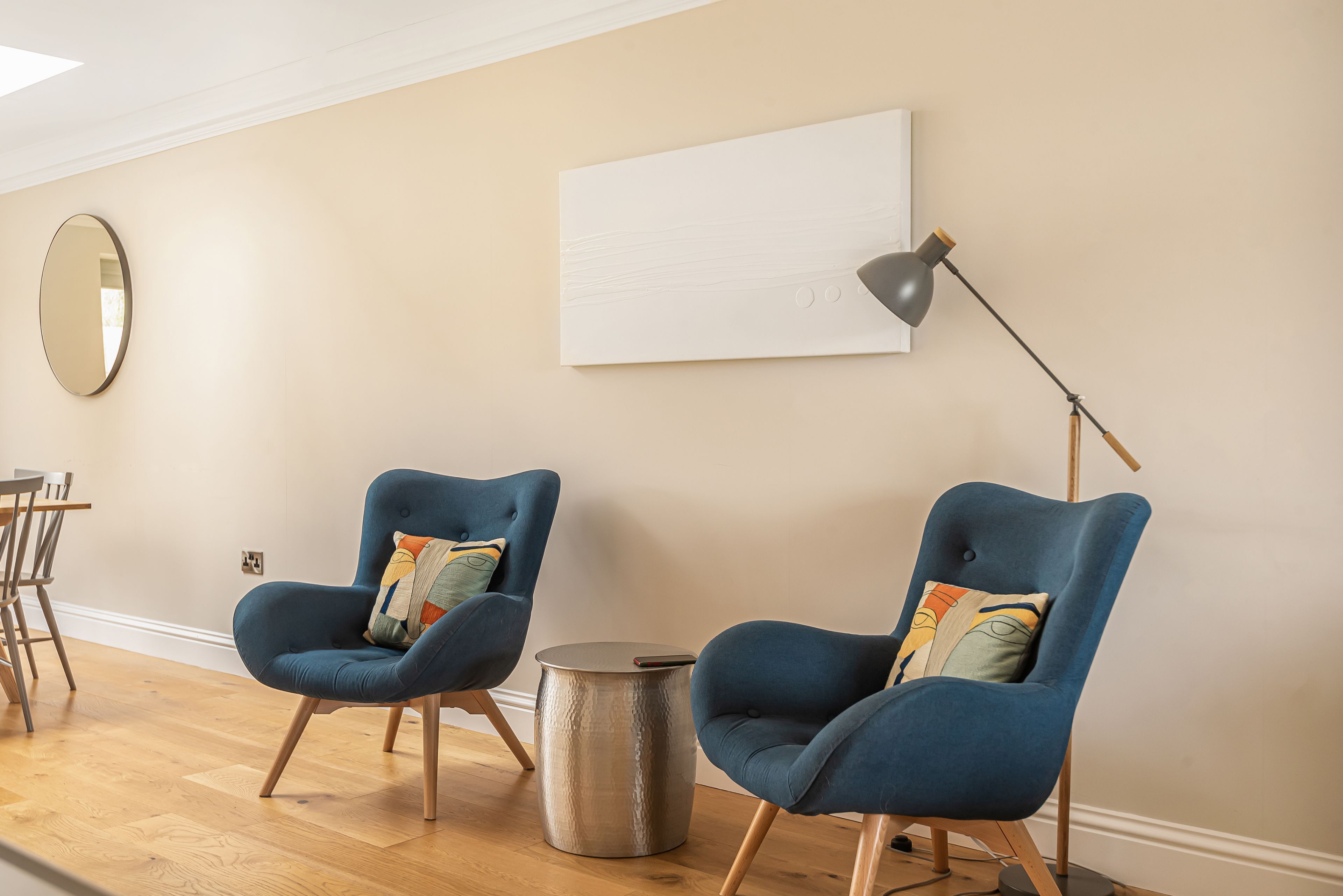 Ein Zimmer mit zwei blauen Sesseln, einem runden Beistelltisch, einer Stehlampe, einem runden Spiegel und einer schlichten Wand.