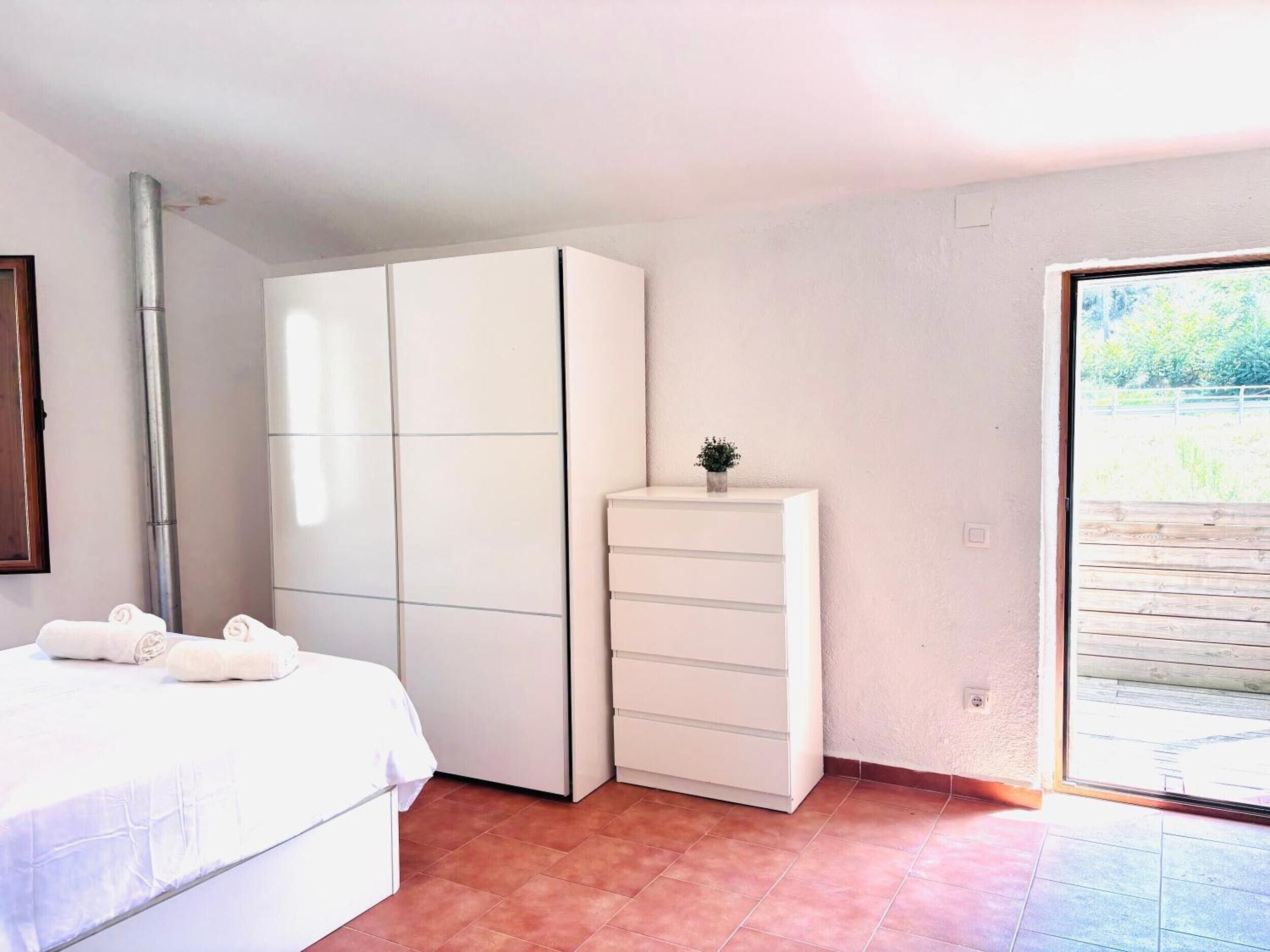 Une chambre à coucher avec un lit, une armoire blanche, une petite plante et une porte-fenêtre coulissante donnant sur un espace extérieur.
