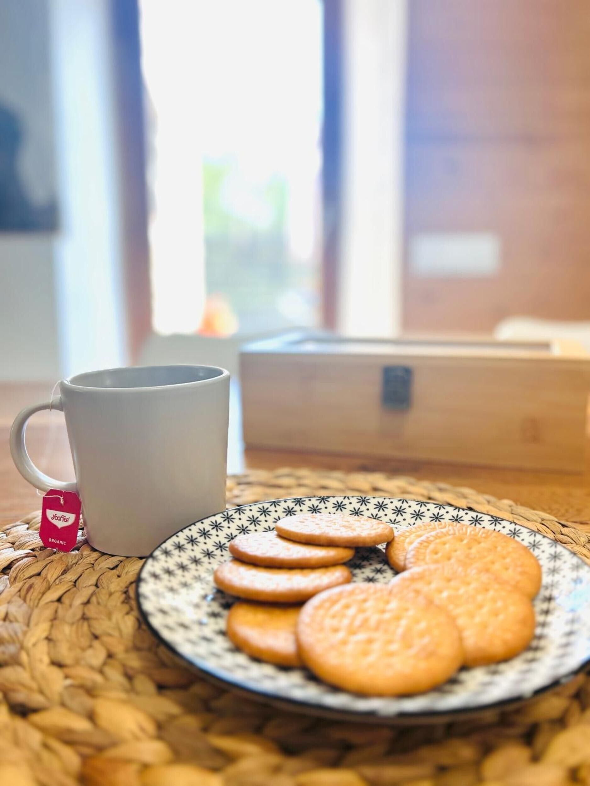 Una taza blanca con una bolsita de té, un plato con galletas y una alfombrilla tejida sobre una mesa.