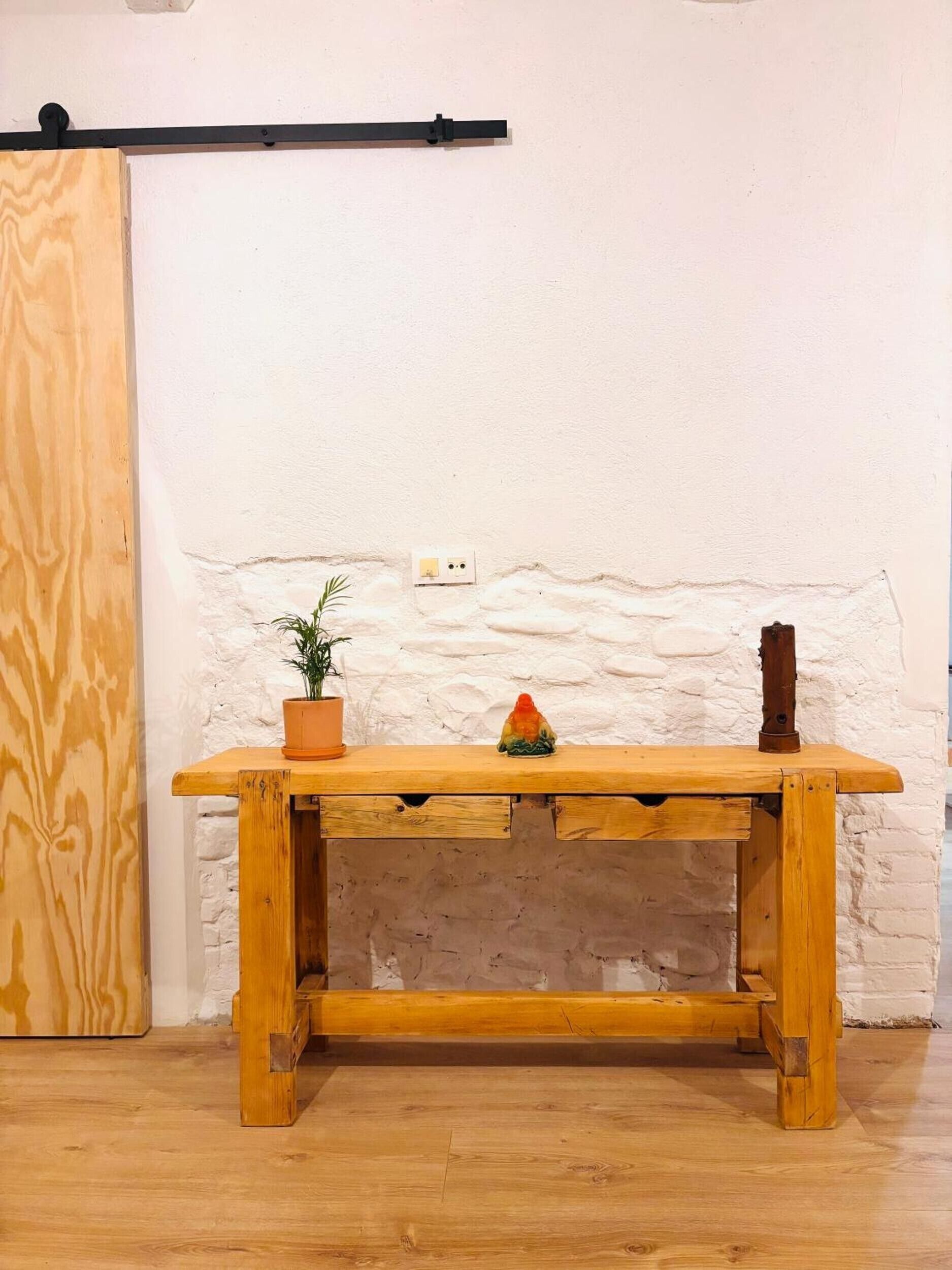 Una mesa de madera con una planta en maceta y un objeto decorativo encima, frente a una pared blanca con textura y una barra horizontal negra arriba.