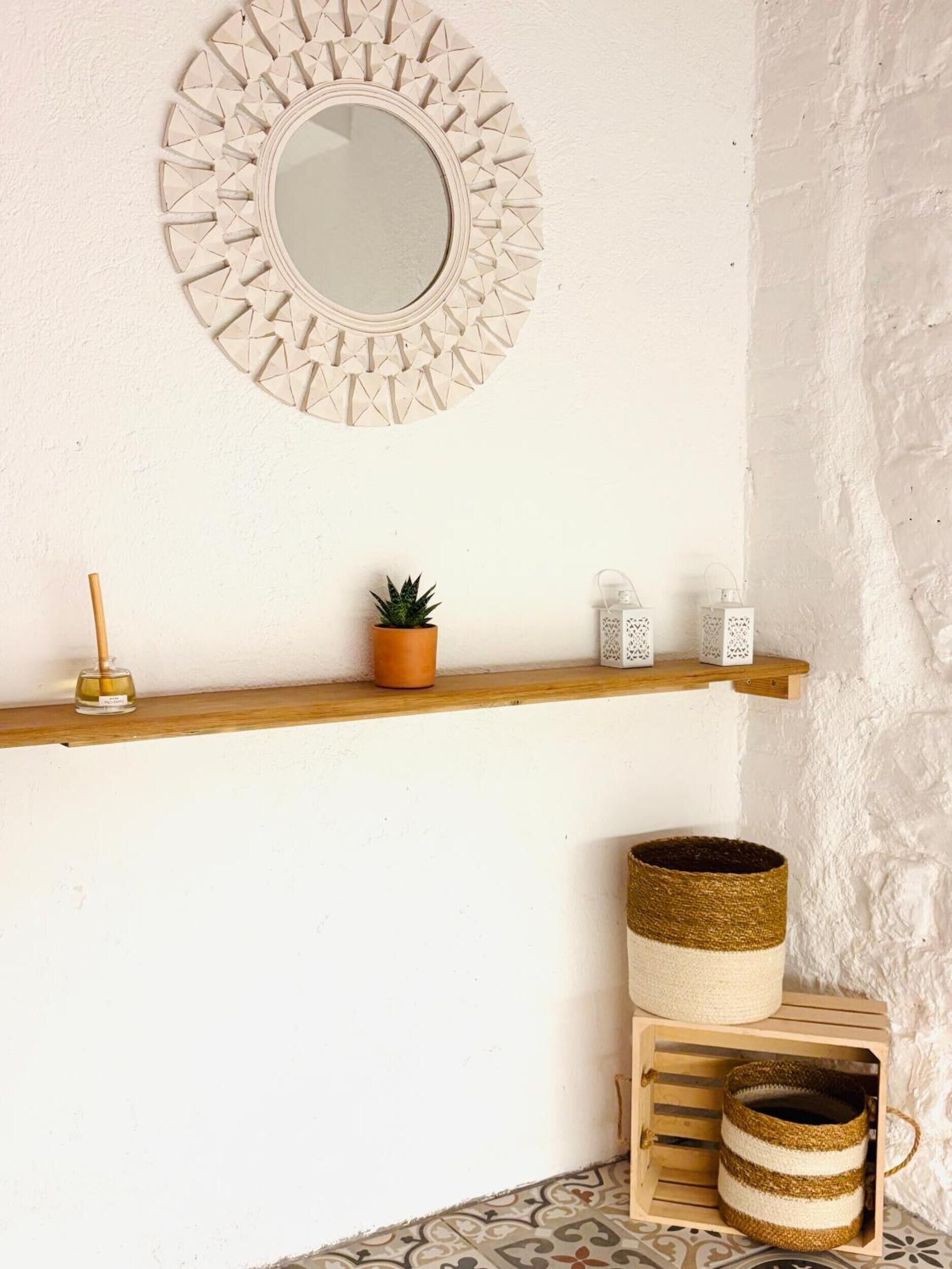Un espejo montado en la pared, un estante con una planta en maceta, dos faroles y una cesta tejida sobre un muro con textura.