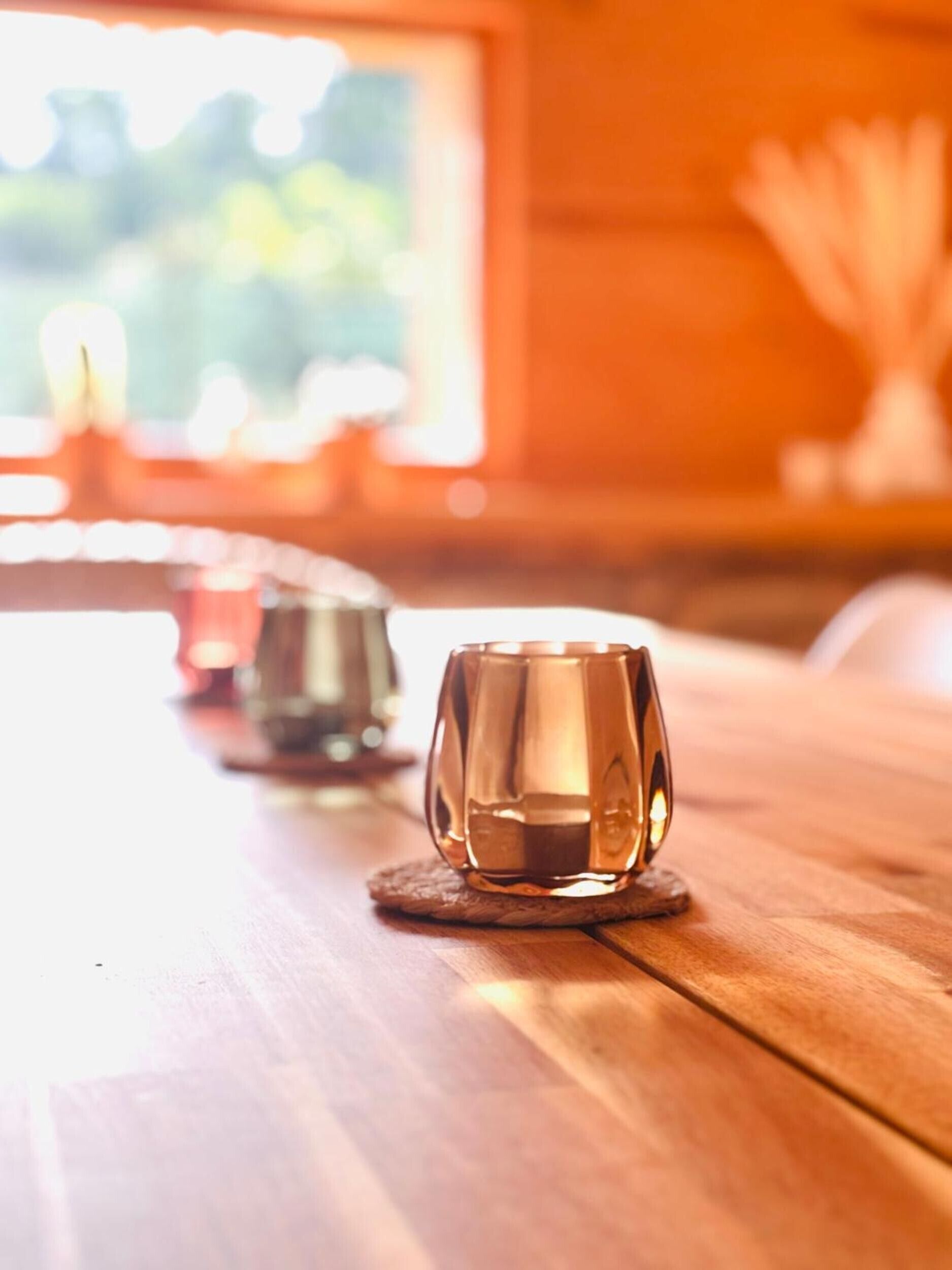 A wooden table with three glass cups on coasters, a window with a view of greenery, and a vase with white flowers.