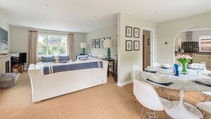 A modern living room with a sofa, a dining table set for six, and a kitchen visible through an archway. - Sanderling - sleeps 7 guests  in 4 bedrooms (Burnham Market)