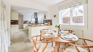 A kitchen with a dining area, featuring a central island, wooden chairs, and a view of a brick building through a window. - Sanderling - sleeps 7 guests  in 4 bedrooms (Burnham Market)
