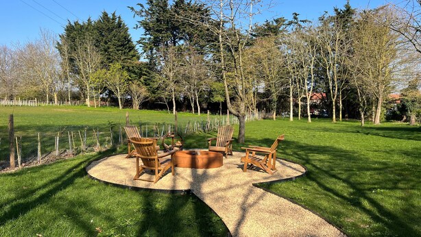A circular outdoor seating area with wooden chairs and a central fire pit, surrounded by a gravel path and a grassy lawn, with trees and a fence in the background. - Sanderling - sleeps 7 guests  in 4 bedrooms (Burnham Market)