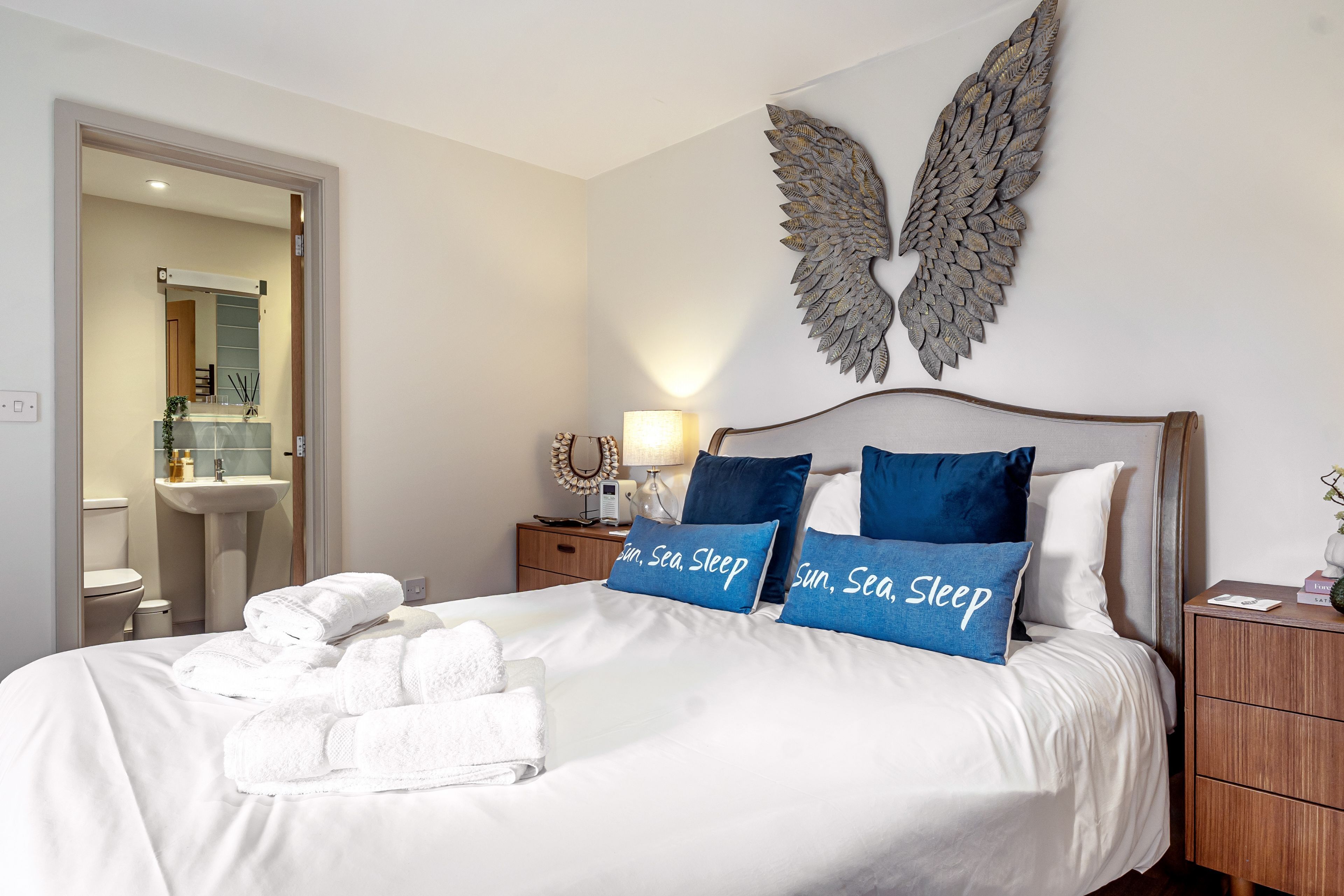 Une chambre d’hôtel avec un lit, une table de chevet, un objet décoratif fixé au mur et une salle de bain visible à travers une porte ouverte.