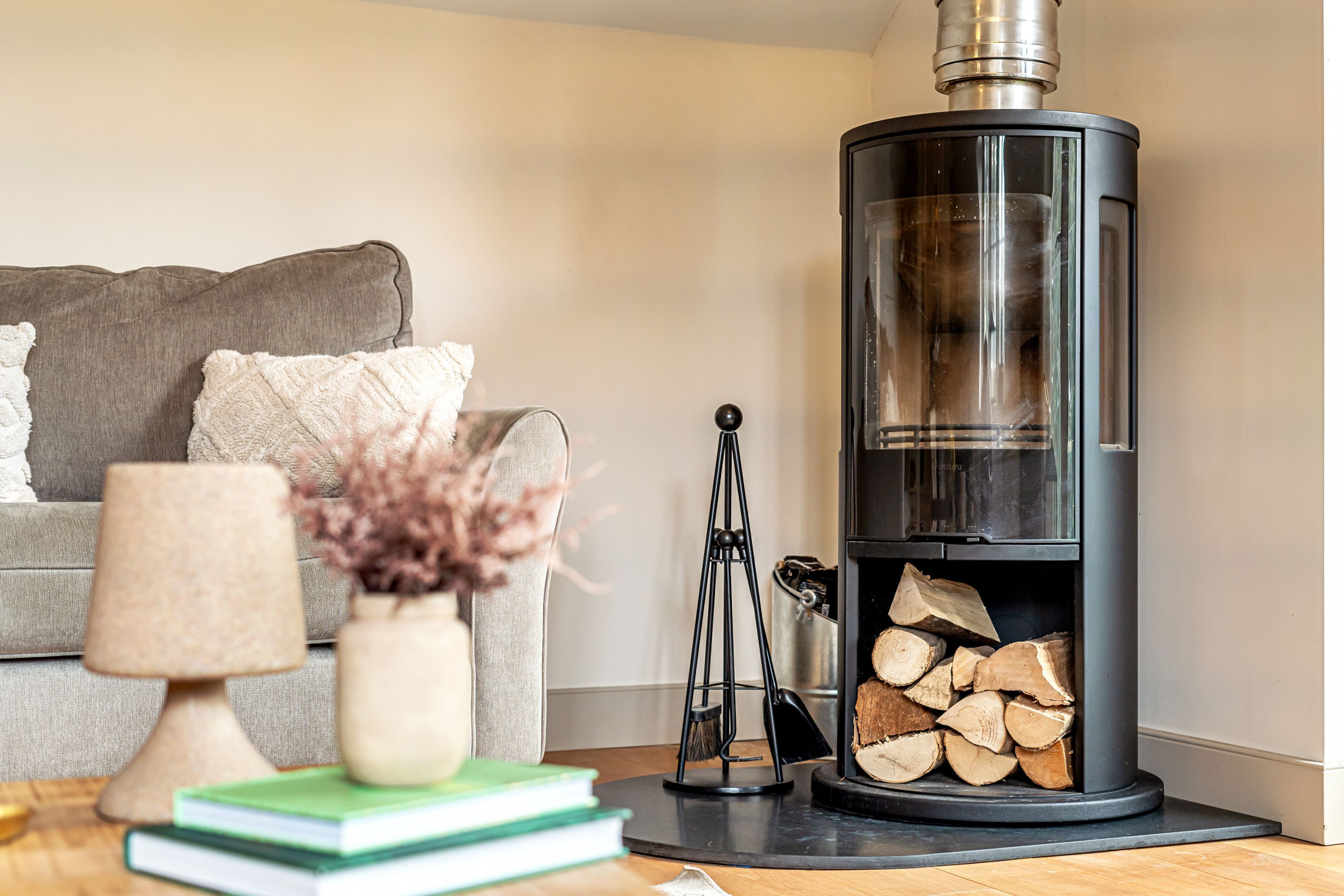 A modern living room with a black wood-burning stove, a grey sofa, a small table with books and a lamp, and a vase with dried flowers.