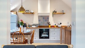 A modern kitchen with white cabinetry, a built-in oven, and a dining area with wooden chairs and a table. - Mill View Burnham Norton - sleeps 4 guests  in 2 bedrooms (Burnham Norton)