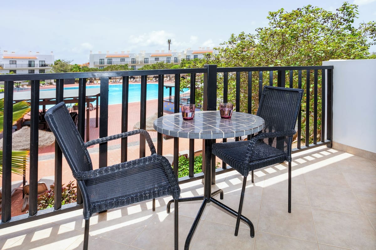 A balcony with a round table and two wicker chairs, overlooking a swimming pool and a building complex.