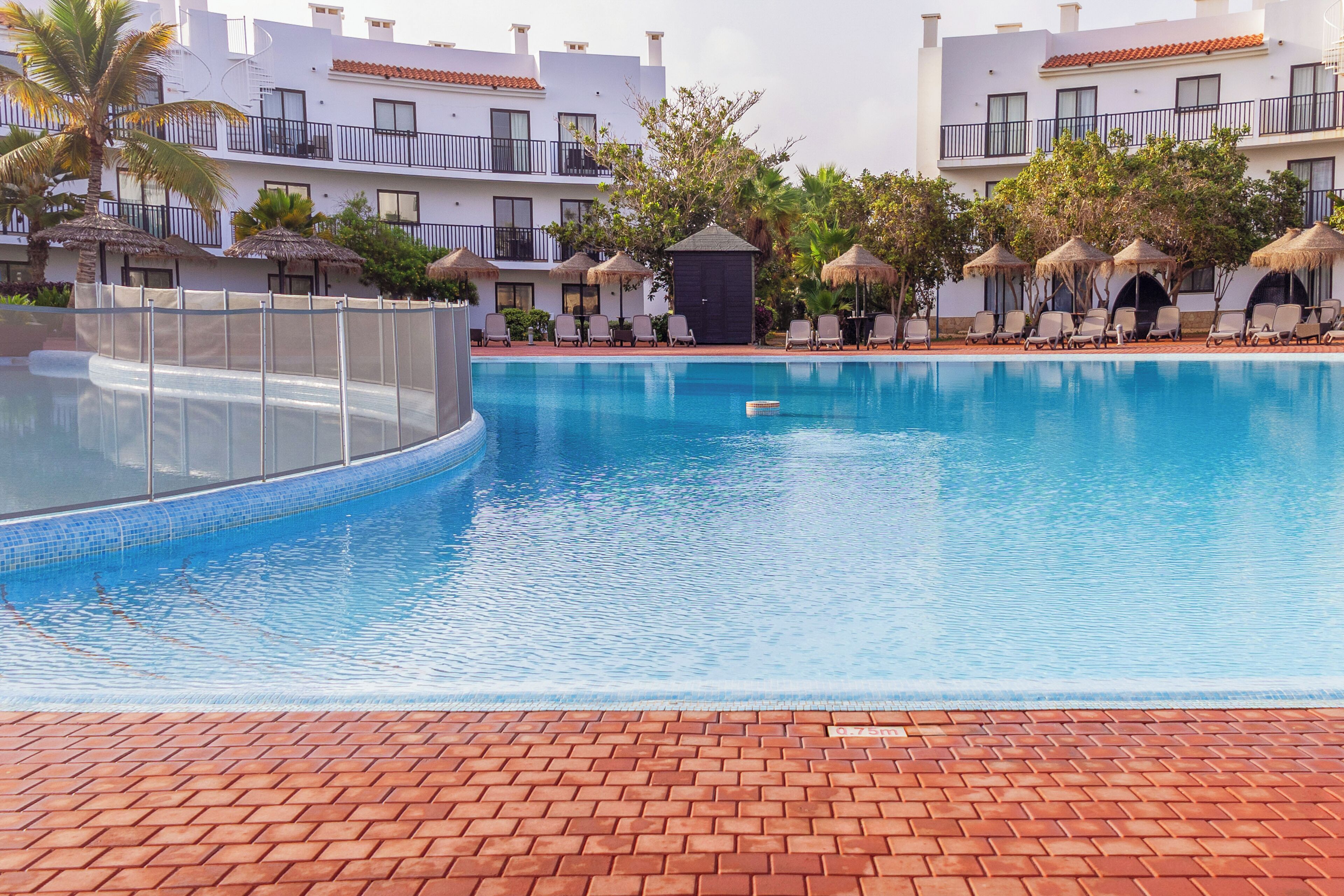 A swimming pool with a brick deck, surrounded by lounge chairs and umbrellas, with a building in the background.