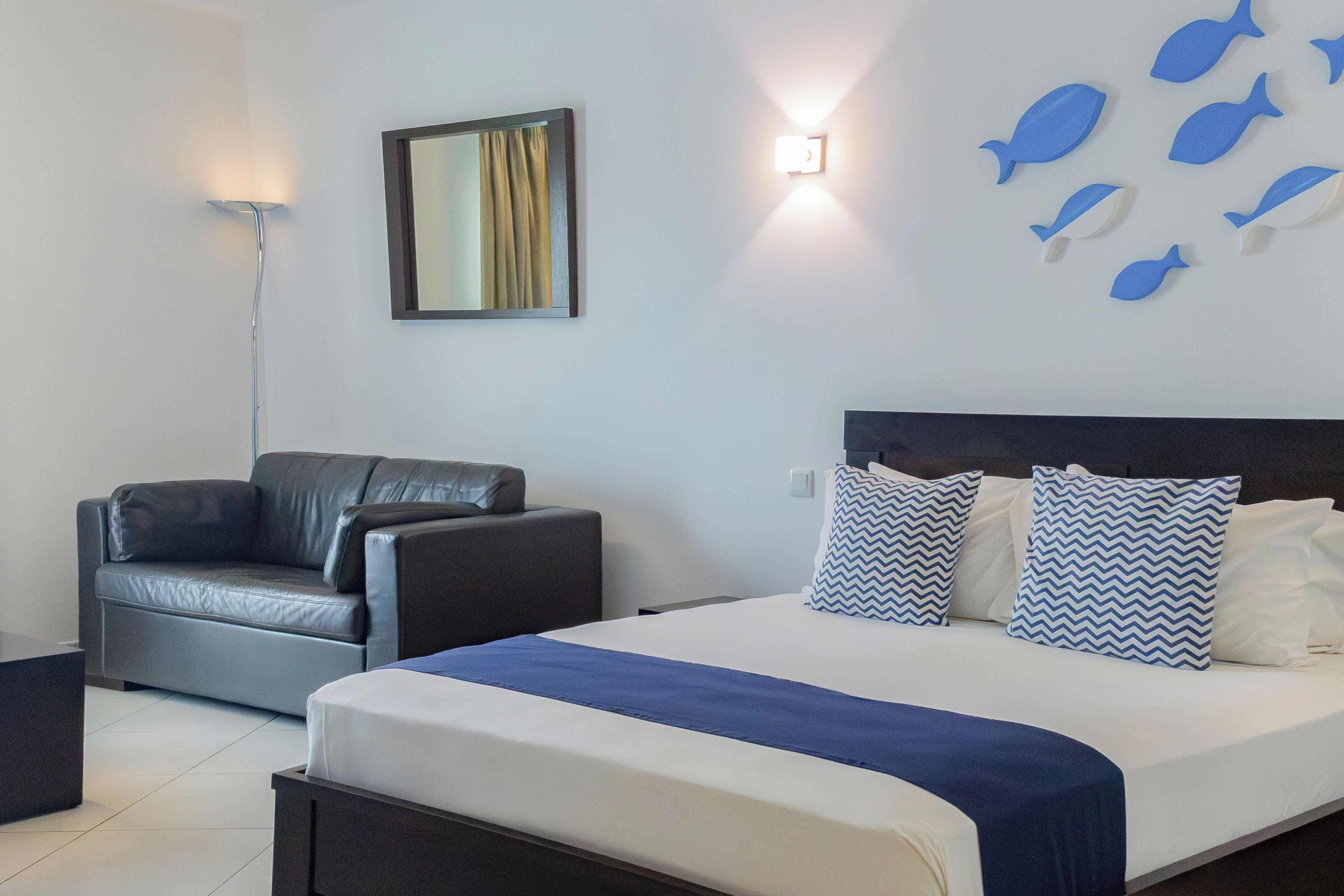 Una habitación de hotel moderna con una cama, un sofá de cuero, un espejo montado en la pared y decoración de peces en la pared.