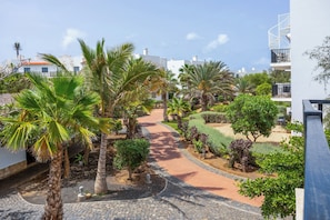 A paved pathway leading through a landscaped area with palm trees and shrubs, surrounded by buildings with balconies.