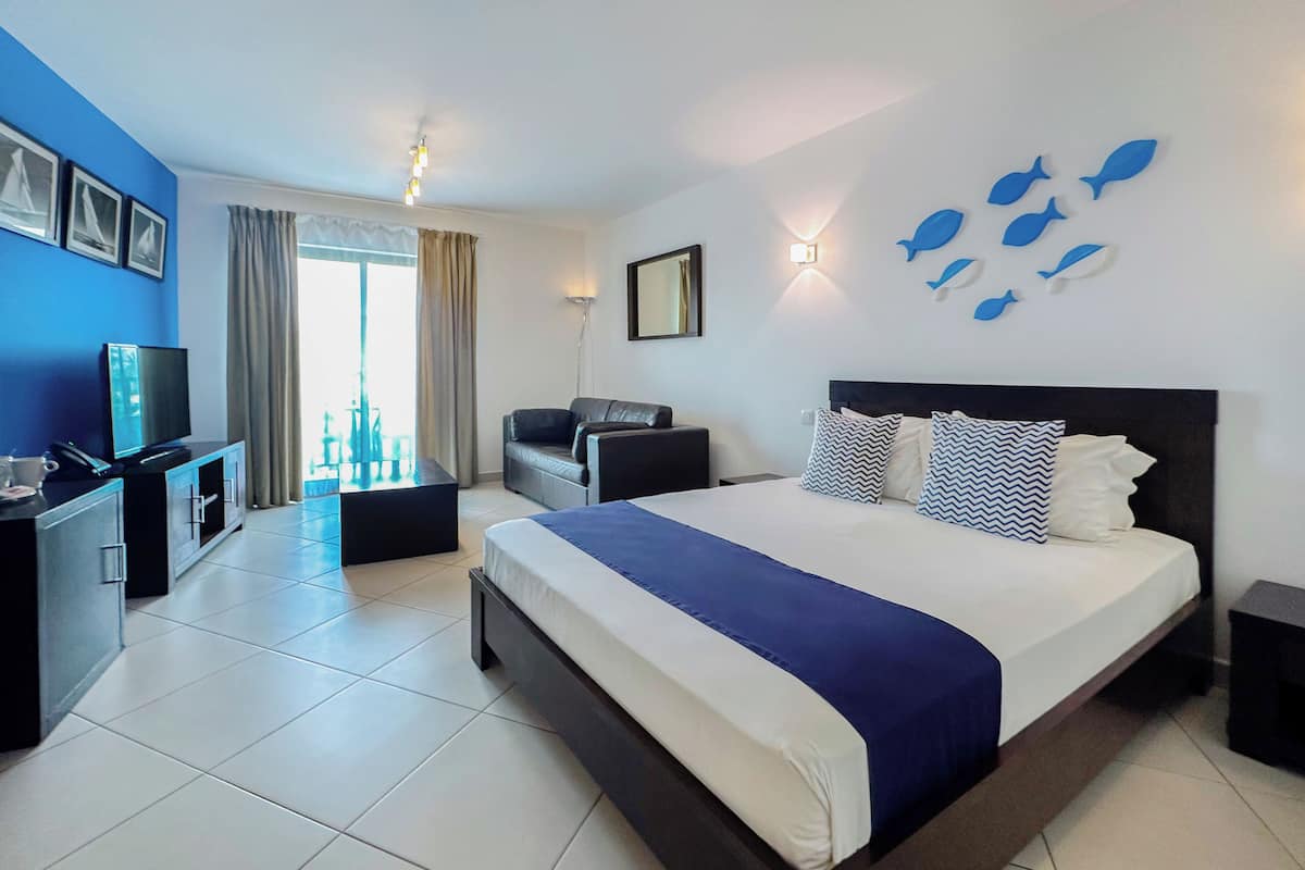 A hotel room with a large bed, a TV, a desk, and a balcony with curtains.