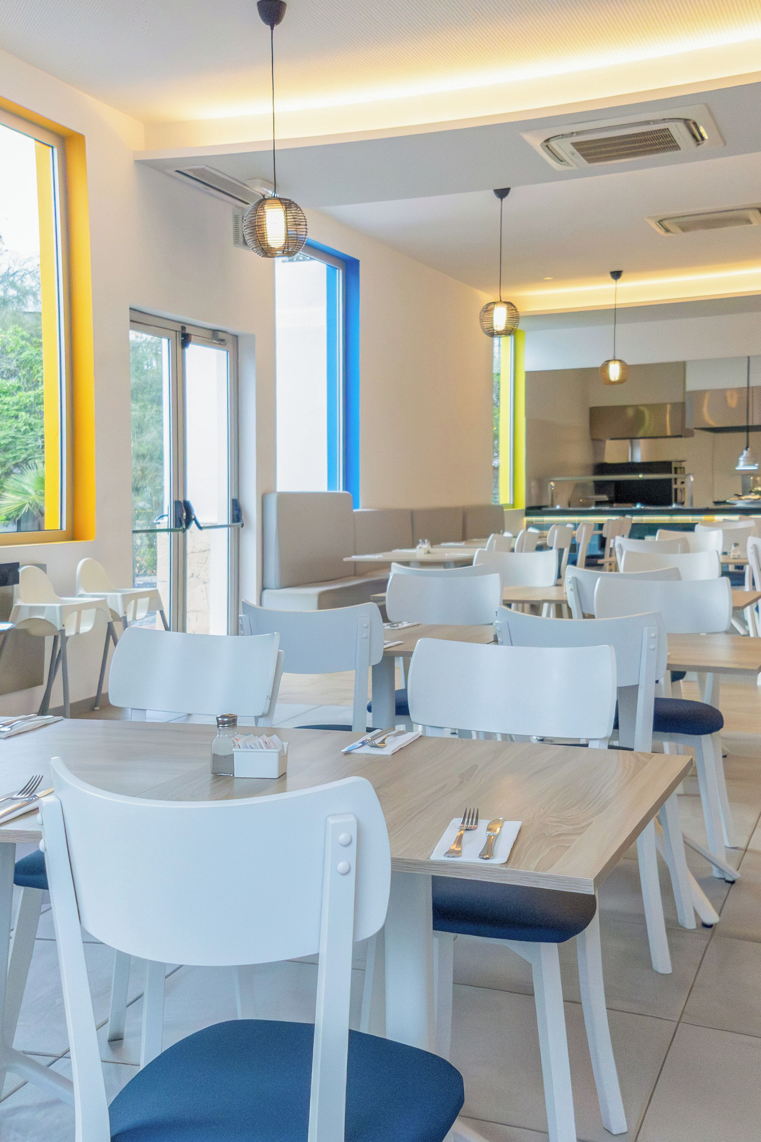 Un espace de restauration moderne avec des chaises et des tables blanches, de grandes fenêtres et une cuisine ouverte visible au fond.