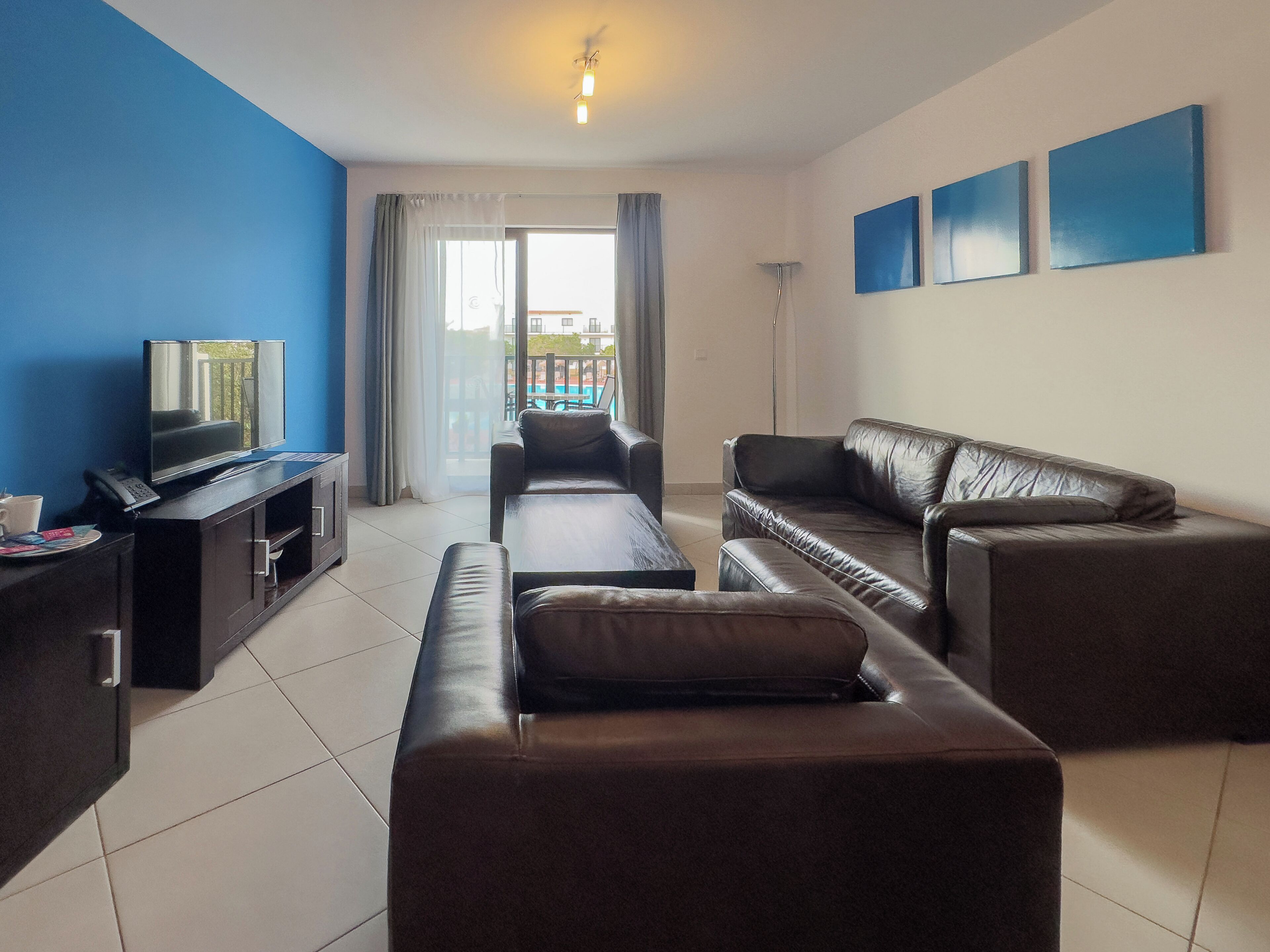 A modern living room with a sofa, a coffee table, and a TV. There is a balcony with a view of buildings and a pool.