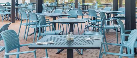 An outdoor patio area with multiple tables and chairs, a covered roof, and a brick floor.