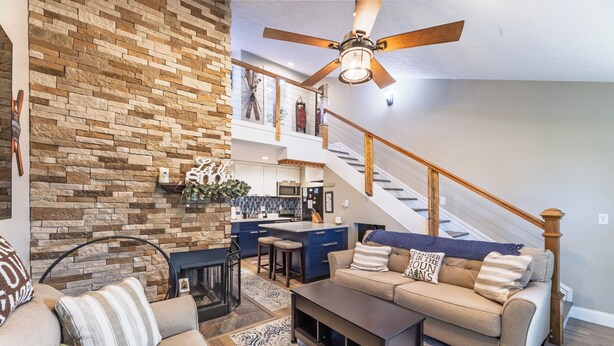 A spacious living room with a stone accent wall, a ceiling fan, a staircase, and a kitchen area. - Cozy 1-bedroom king condo w/fireplace in Snowshoe. Walk to slopes! (Snowshoe)