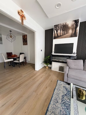Un salon moderne doté d’un téléviseur à écran plat, d’un canapé gris, d’un coin repas avec une table et des chaises en bois, et d’un couloir menant à une autre pièce.
