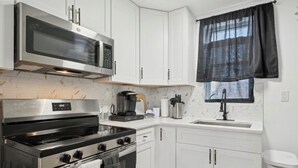A compact kitchen with white cabinets, a stainless steel oven, a microwave, a coffee maker, and a window with a black curtain.