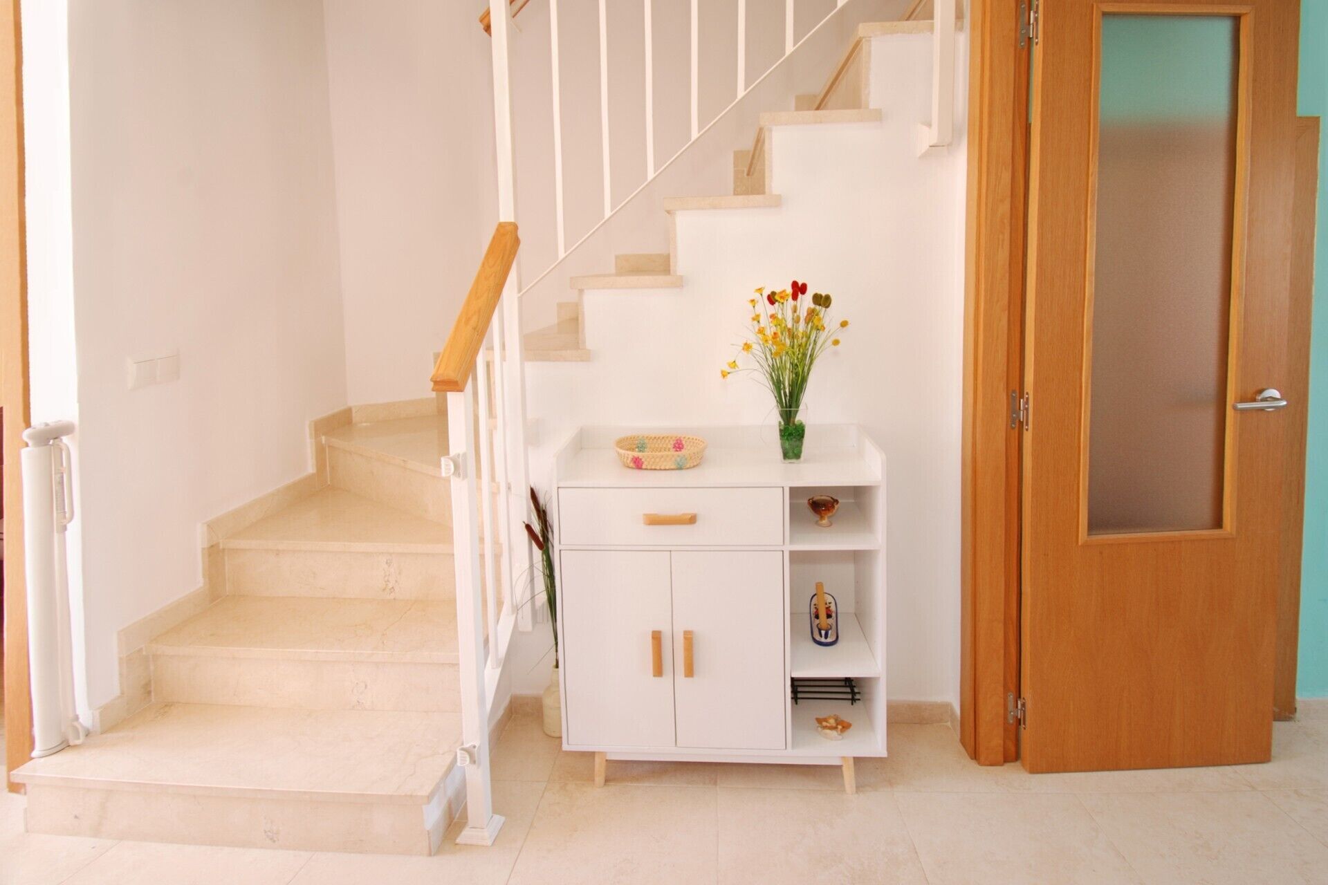 Una escalera con barandilla blanca, un armario blanco con cajones y estantes, un florero con flores y una puerta de madera.