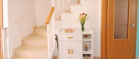 Una escalera con barandilla blanca, un armario blanco con cajones y estantes, un florero con flores y una puerta de madera.