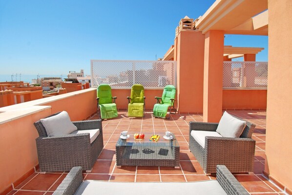 A rooftop terrace with wicker furniture, a glass coffee table, and a view of the cityscape.