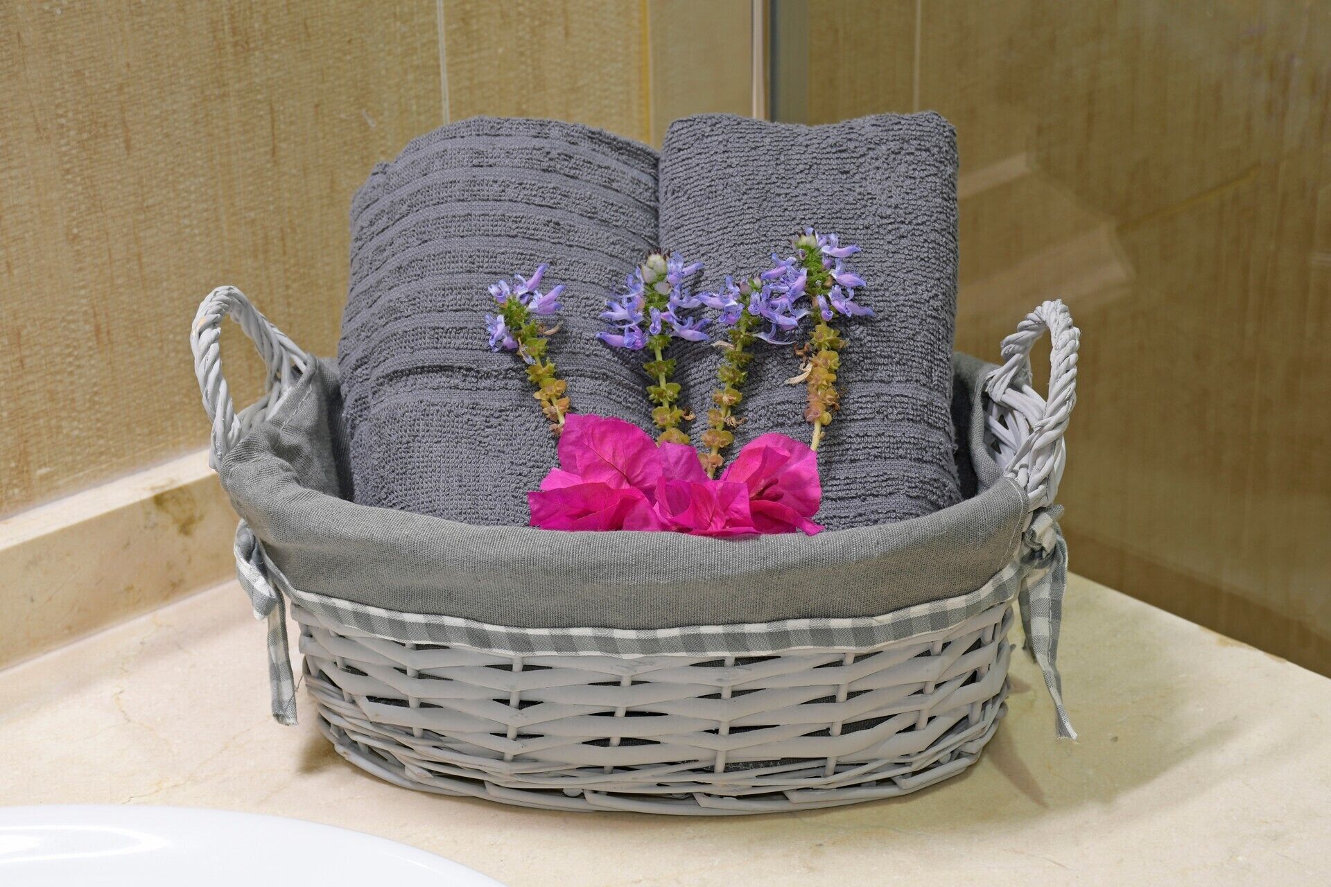 A wicker basket with two gray towels and purple flowers on a bathroom counter.
