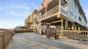 Una terraza de madera con una mesa de picnic y barandilla, con vista a una playa y un edificio con balcones.