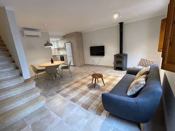 Een moderne woonkamer met een bank, een eethoek met stoelen, een aan de muur bevestigde televisie en een open haard.